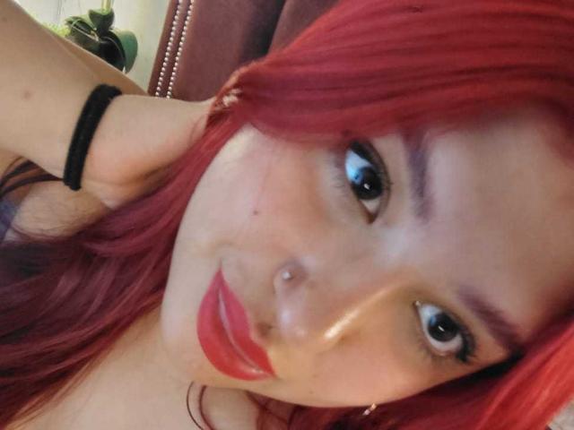Selfie de primer plano de AmberCutel con cabello rojo vibrante, labios rojos grandes y ojos oscuros. Tiene una goma para el pelo negra en la muñeca y un piercing en la nariz. Expresión sensual, con una ligera sonrisa..