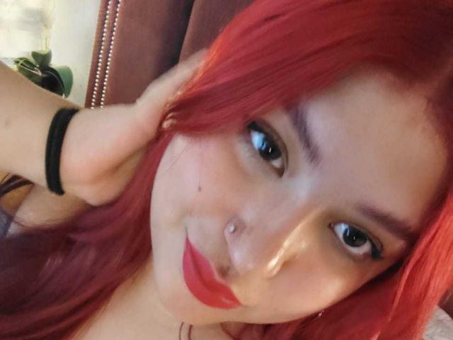 Fotografia ravvicinata di AmberCutel con capelli rossi, pelle chiara, sopracciglia nere, piercing al naso e rossetto rosso. Lei sta guardando la fotocamera con una mano che le sfiora i capelli. Molto attraente..