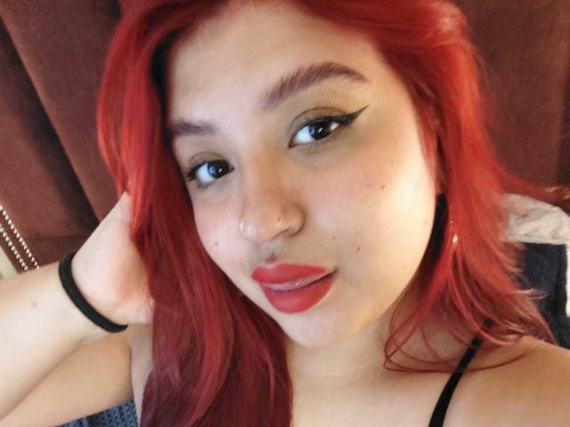 Primo piano di AmberCutel con capelli rossi vivaci, eyeliner nero spesso e rossetto rosso. Lei guarda la fotocamera con un leggero sorriso, e la sua mano è appoggiata sulla testa. Molto attraente..