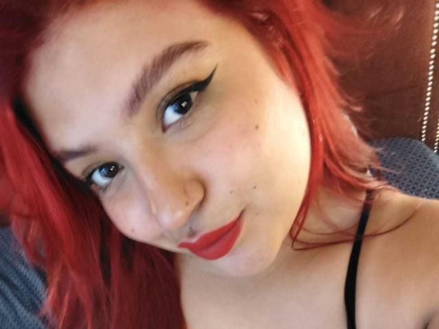 Gros plan d'AmberCutel avec de magnifiques cheveux roux, un trait d'eye-liner noir et du rouge à lèvres rouge. Elle se penche en avant, établissant un contact visuel direct avec une bouche légèrement entrouverte, dégageant une tension sexuelle. Peau charnue, pâle..