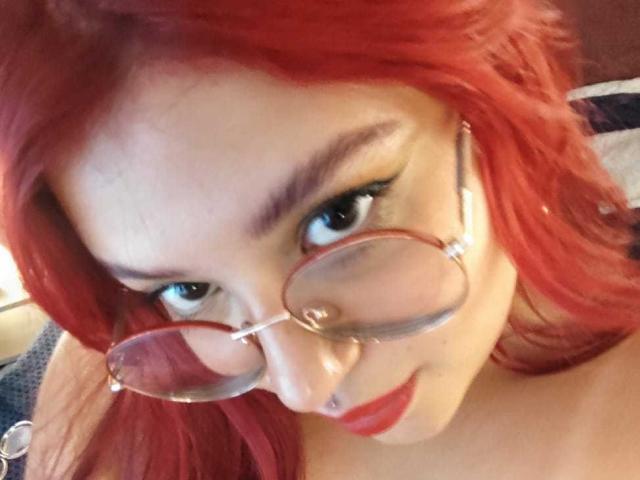 Close-up foto van AmberCutel met levendig rood haar, die ronde brillen draagt, omhoog kijkt met intense blauwe ogen, en rood lippen.

zichtbare decolleté suggereert naaktheid, wat een seksuele spanning creëert..