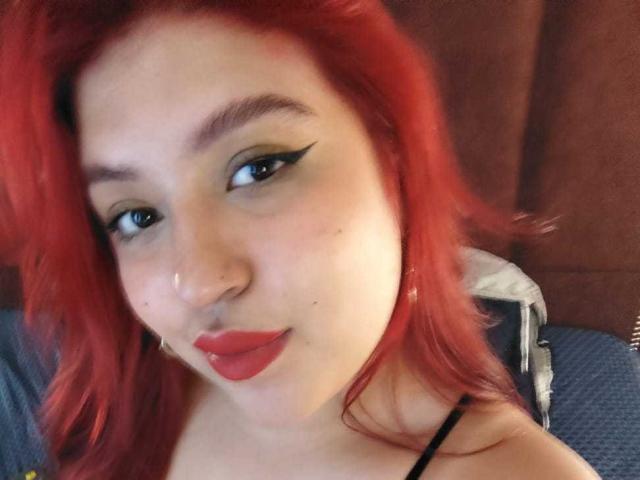 Foto em close-up de AmberCutel com cabelo ruivo vibrante, delineador preto marcante e batom vermelho. Ela está olhando diretamente para a câmera com um leve sorriso, exibindo seus lábios cheios e pele clara..