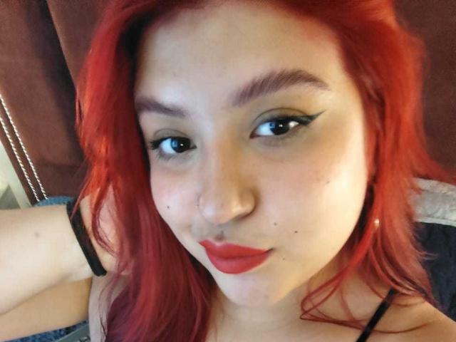 Primer plano de AmberCutel con cabello rojo, piel pálida y delineador de ojos negro. Ella lleva pintalabios rojo, mirando directamente a la cámara con una ligera sonrisa. Correa visible de un sujetador negro. Tensión sexual.