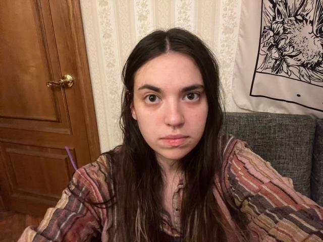 Close-up of LenoreSaile, a young woman with long dark hair, wearing a striped shirt, looking directly at the camera with a serious expression. Wooden door and wall art in background.