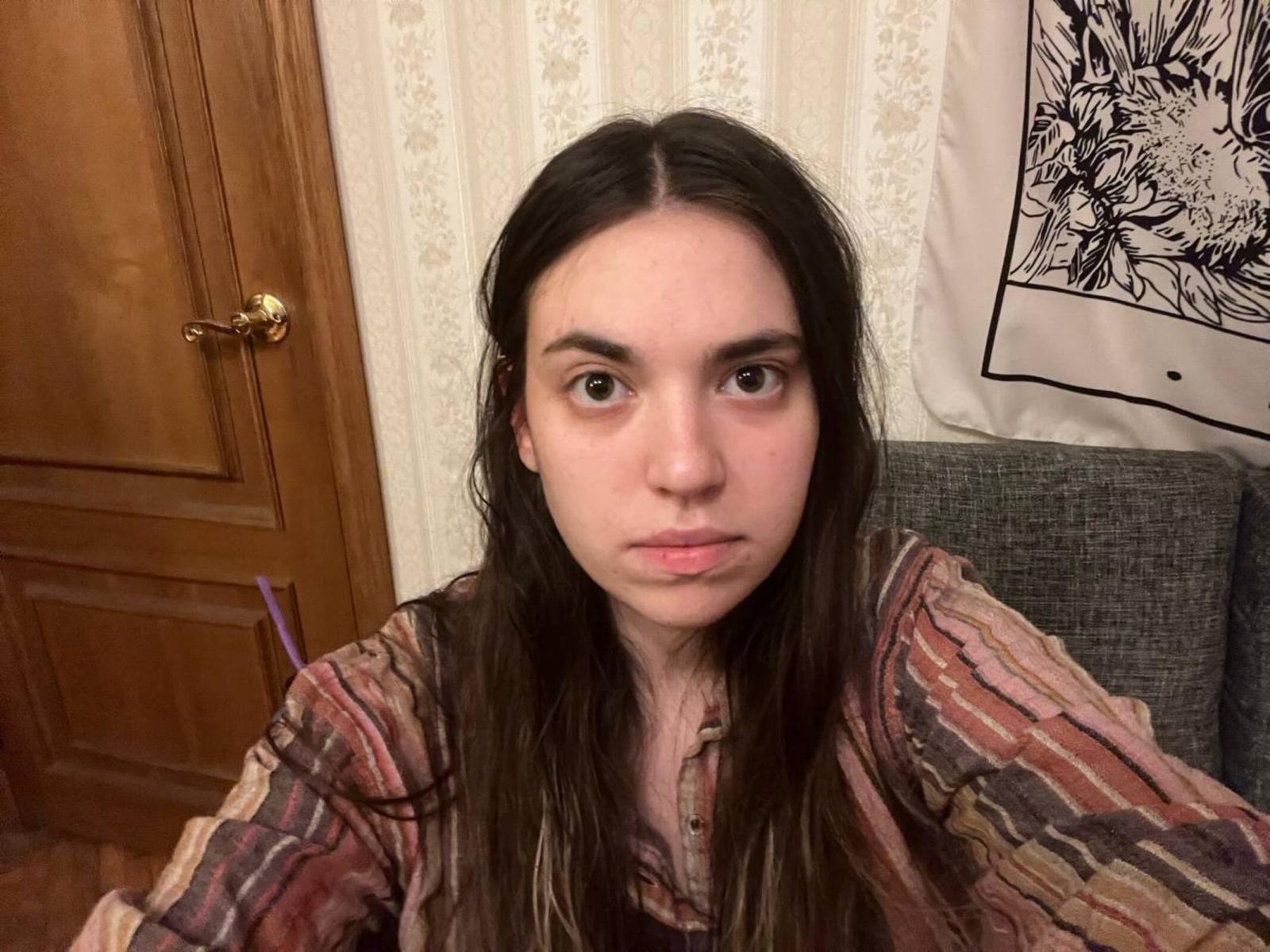 Primo piano di LenoreSaile, una giovane donna con lunghi capelli scuri, che indossa una camicia a righe e guarda direttamente la fotocamera. Un'espressione seria. Porta in legno e decorazioni murali sullo sfondo..