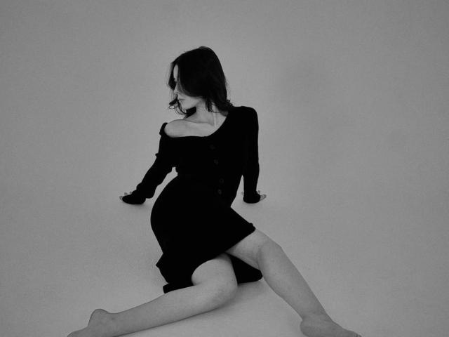 TrinidadGuerini sitzt auf einem grauen Boden, die Beine gespreizt, trägt ein schwarzes Kleid mit einem nackten Schulterteil und blickt mit dunklem Haar nach unten. teilweise bedecktes Gesicht. Monochromer blau-grauer Filter, verführerpose, leichter Schatten unter dem Körper..
