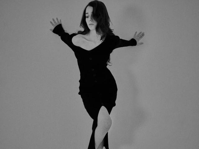 Photograph of TrinidadGuerini with long dark hair, leaning against a wall, arms spread wide. Wearing a black off-shoulder dress, accentuating large breasts. Shadowed, moody, monochromatic blue lighting. Sexy, provocative pose.