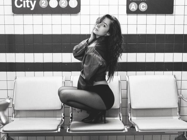 Black and white photo of TrinidadGuerini squatting on a subway bench, wearing a denim jacket and black shorts, hands near face, looking seductively to the side. Bright tiled wall background.