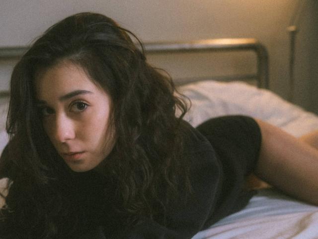 TrinidadGuerini è sdraiata su un letto, a faccia in giù, con i capelli scuri e ondulati e occhi intensi. Indossa una maglia nera che mette in risalto le sue forme e le sue gambe. Una luce soffusa illumina la sua pelle..