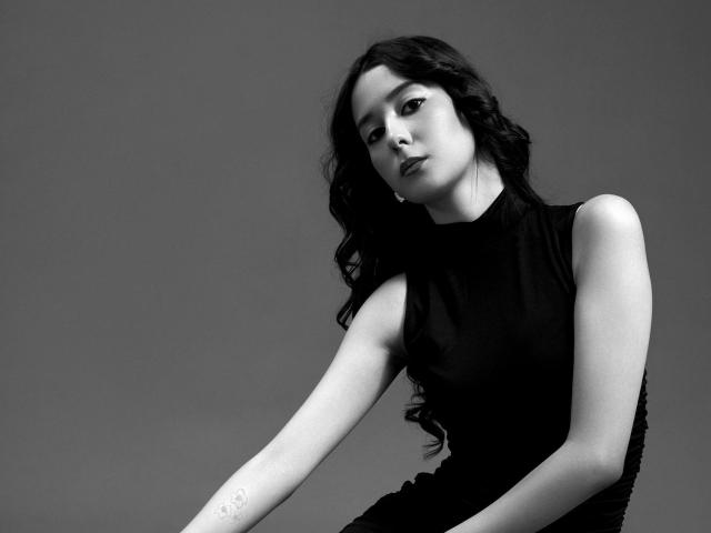 Black and white photograph of TrinidadGuerini with long, wavy hair, wearing a sleeveless black top, leaning forward, arm extended, intense expression, high contrast, sharp shadows, sensual tension, monochrome.