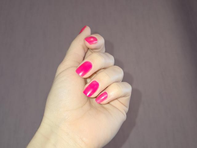 Primer plano de la mano de Amberia, con esmalte de uñas rosa brillante, los dedos ligeramente curvados, mostrando uñas largas y brillantes. La iluminación suave resalta la textura de la piel. El fondo es una superficie lisa y de color gris oscuro..