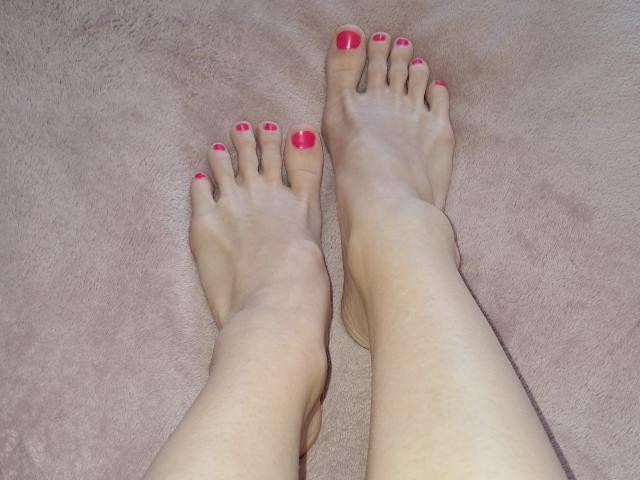 Los pies descalzos de Amberia, con las uñas de los pies de un rojo brillante, las piernas ligeramente separadas, los dedos apuntando ligeramente hacia afuera, sobre un beige texturizado. alfombra. El tono de piel es claro, los pies parecen suaves y bien cuidados..