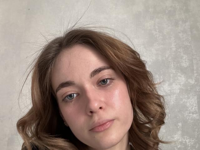 Photographie en gros plan de CelestineSegalla, une jeune femme à la peau claire, aux yeux bleus et aux cheveux bruns ondulés. Elle a une expression neutre, des lèvres rosées et un visage légèrement rougi. Le fond est une surface blanche texturée..