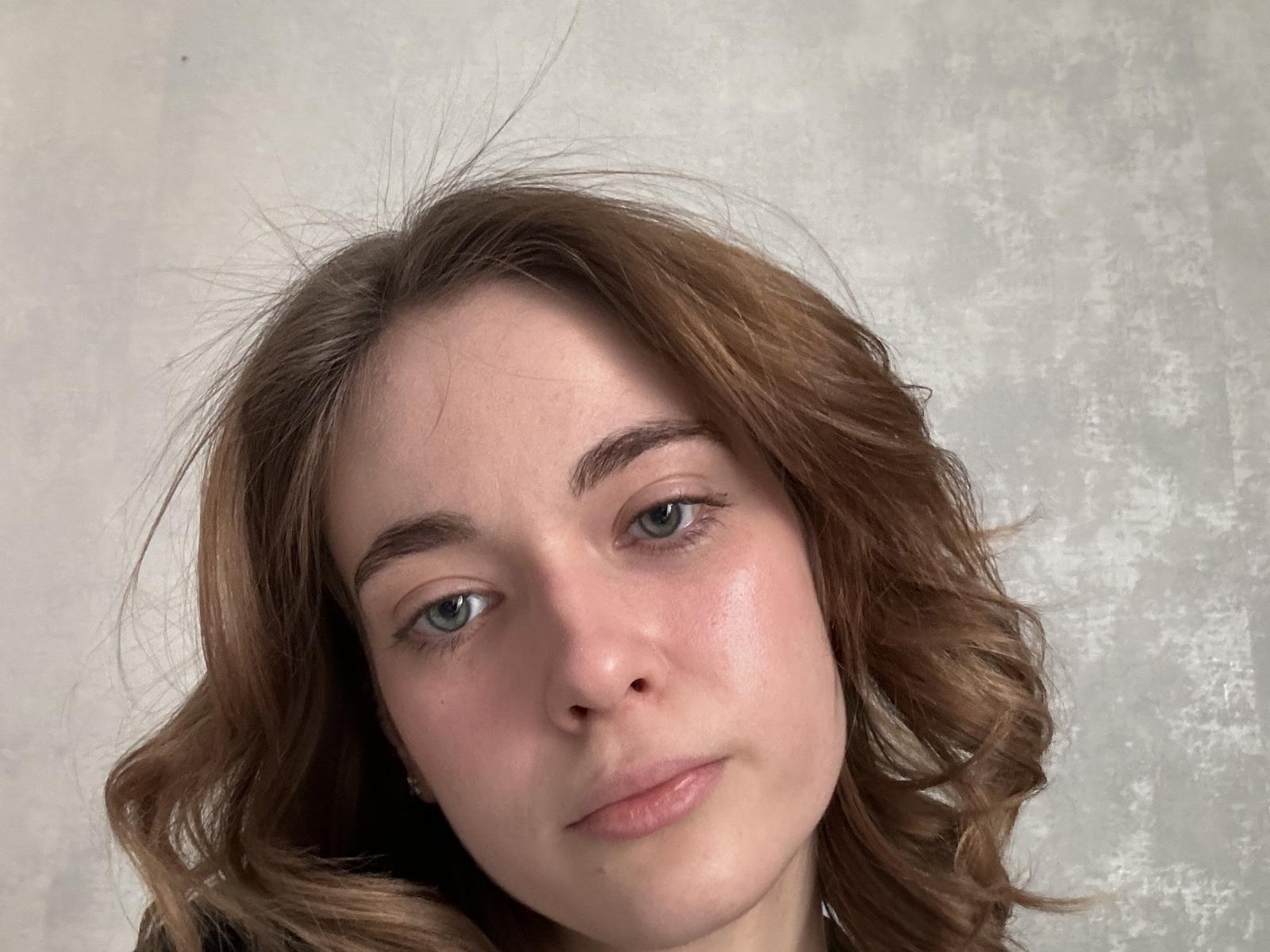 Close-up photograph of CelestineSegalla, a young woman with fair skin, blue eyes, and wavy brown hair. She has a neutral expression, soft pink lips, and a slightly flushed face. Background is a textured white surface.