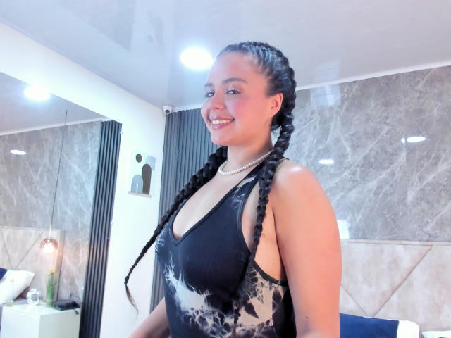 ArielleWilde with braided hair, smiling, wearing a black lace-up halter top, pearl necklace, in a modern room with marble walls. Bright lighting, confident pose, sexy vibe, attractive.