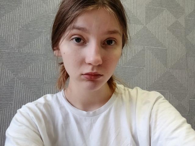 Photographie de Peggy Ricley avec des cheveux bruns lisses, portant une chemise blanche, face à l'appareil photo, avec une expression neutre. L'arrière-plan présente un motif géométrique texturé. Une tension sexuelle est visible, due au regard direct..