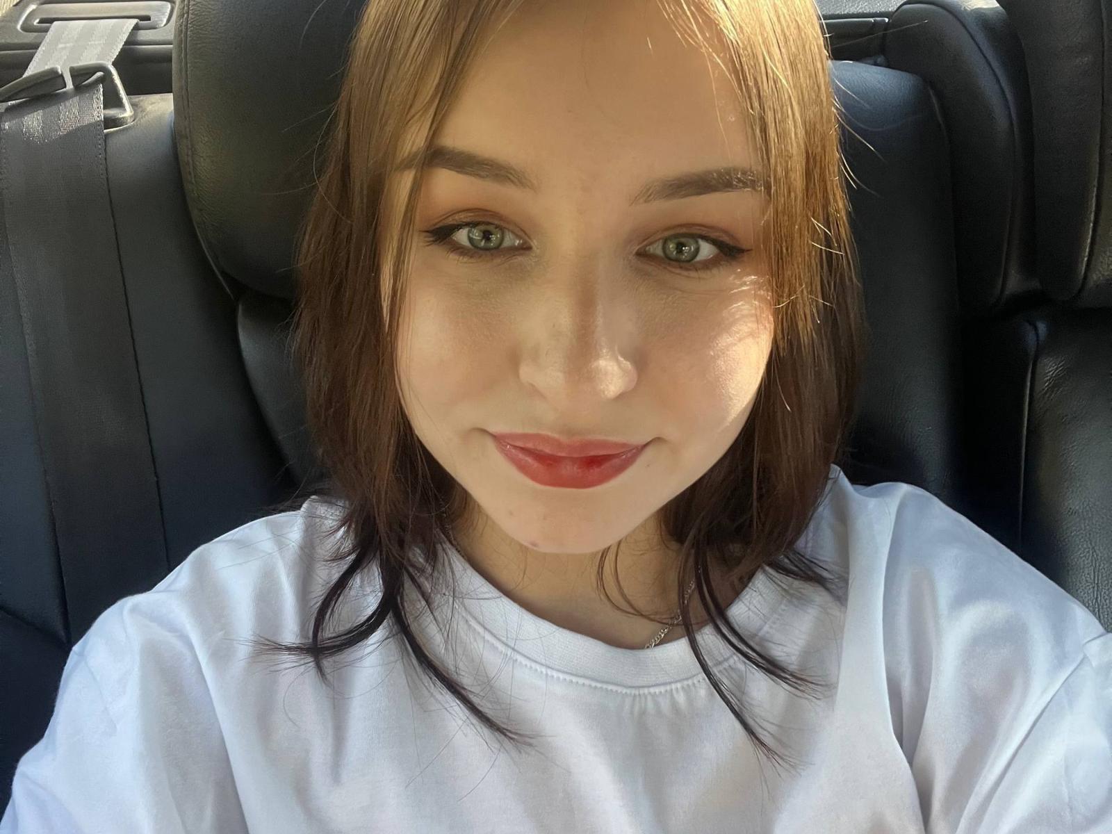 Close-up selfie van Etta Prentice in een autostoeltje, gekleed in een witte blouse, met lichtbruin haar, groene ogen en rood lipsticks. Het zonlicht accentueert haar gezicht, waardoor ze een sensuele, verleidelijke uitstraling krijgt..