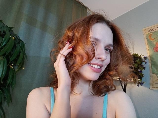 LourieHegner heeft rood, golvend haar, ze glimlacht met zichtbare tanden en raakt haar haar aan met één hand. Ze draagt een lichtblauwe top met bandjes. Op de achtergrond staan een groene plant en een poster met een kaart. Heel schattig.