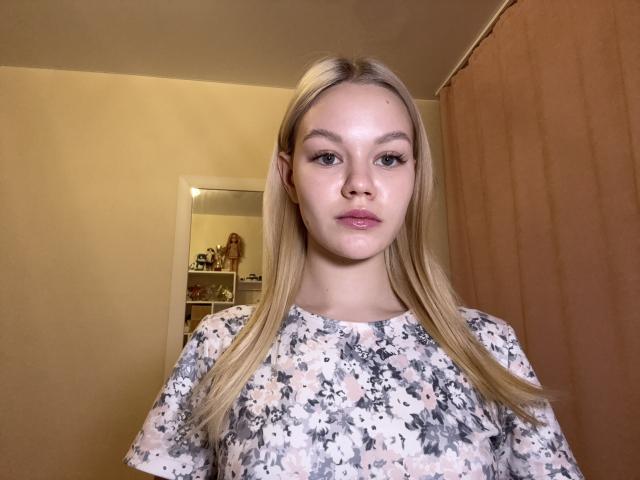 TonyEppihimer heeft lang blond haar, een lichte huid en draagt een bloemenjurk. Ze kijkt recht in de camera met een neutrale uitdrukking en staat in een kamer met een spiegel..