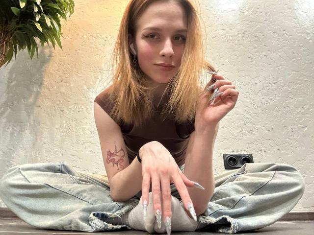 Lourie Hegner è seduta a gambe incrociate sul pavimento, indossa una maglietta marrone e jeans strappati, e tiene una sigaretta nella mano destra. Ha i capelli lunghi e biondi e un tatuaggio sul braccio sinistro..