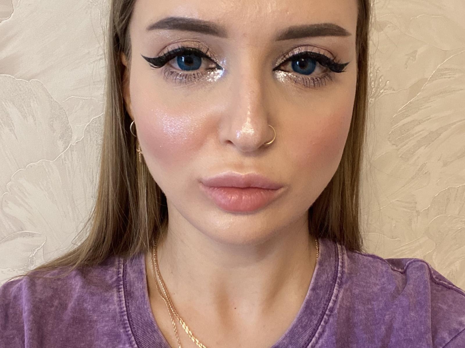 Photo en gros plan de BulaKrugman, une jeune femme aux yeux bleus, avec un trait d'eye-liner noir épais, des lèvres roses brillantes et un petit anneau de nez. et de longs cheveux bruns. Portant une chemise violette, peau claire. Absolument magnifique..