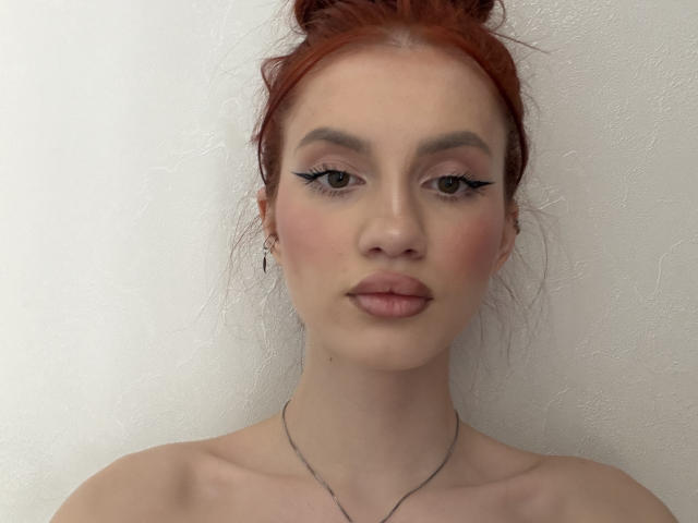 Nahaufnahme von HolleyMeholick, mit heller Haut und roten Haaren zu einem unordentlichen Dutt gebunden, mit schwarzem Eyeliner und rosa Lippenstift. freie Schultern, zarte Halskette am Schlüsselbein, weißer Hintergrund, leicht verführerischer Blick.