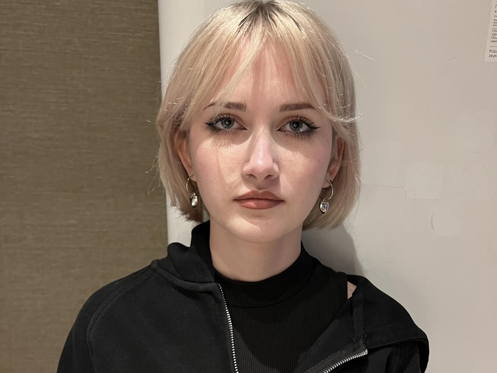 Fotografía de Frances Belfort, con un corte de pelo rubio corto, piel pálida, sudadera con capucha negra, expresión seria, ligera sombra en el rostro. Fondo blanco, iluminación suave, perforación de oreja visible, sutil tensión sexual en la mirada directa..