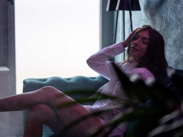 MiyaStar, con lunghi capelli castani, indossa una camicia bianca ed è sdraiata su un divano verde, con le gambe divaricate e una mano tra i capelli, con un sorriso luminoso. luce dietro di lei, ombre scure, forte tensione sessuale.