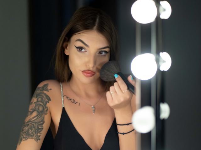 MiyaStar, een donkerharige vrouw met een getatoeëerde arm, brengt foundation aan met een kwastje, voor een rij van felle lampen. Ze draagt een zwarte top met een lage halslijn, rode lippenstift en een ketting. Heel aantrekkelijk..