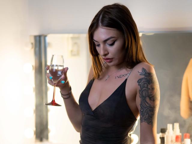 MiyaStar, met donkerbruin haar, draagt een zwarte jurk met een lage halslijn en houdt een martini-glas vast met een rood-oranje drankje. Getatoeëerde arm, verleidelijke blik, zacht licht, subtiele schaduwen. Heel aantrekkelijk.