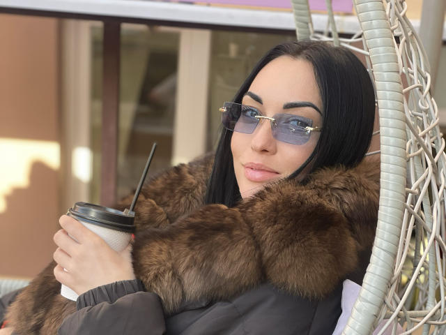 Photograph of Xioni with long black hair, wearing blue reflective sunglasses and a fur-lined black coat, holding a coffee cup with a black straw. Slight sexual tension, confident expression.