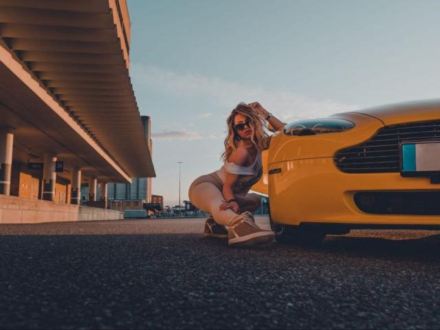 AlmaVelia, agenouillée sur l'asphalte à côté d'une voiture jaune, portant des lunettes de soleil, un débardeur blanc et un short beige, avec un bronzage. bottes.
la lumière du soleil projette de longues ombres, créant une forte tension sexuelle. Cadre moderne, urbain..