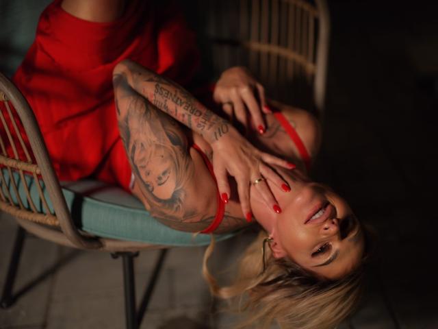 AlmaVelia è sdraiata sulla schiena su una sedia, indossa un vestito rosso, le sue mani con le unghie rosse toccano il suo viso, il suo braccio tatuato. visibile.
capelli biondi, sfondo scuro, posa seducente.