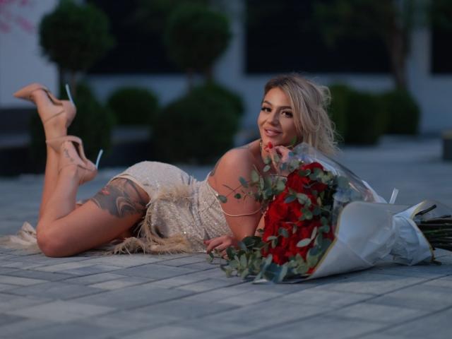 AlmaVelia, una donna bionda con tatuaggi, sdraiata su un lato su un pavimento piastrellato, indossa un abito bianco in pizzo, tacchi alti. tenendo un bouquet di rose rosse, sorriso seducente, illuminazione soffusa.