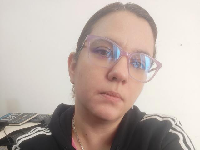 Close-up of AmeliaWilson, wearing clear glasses, black tracksuit with white stripes, and a thin gold necklace. She has light skin, brown hair pulled back, and a serious expression. Bright background.