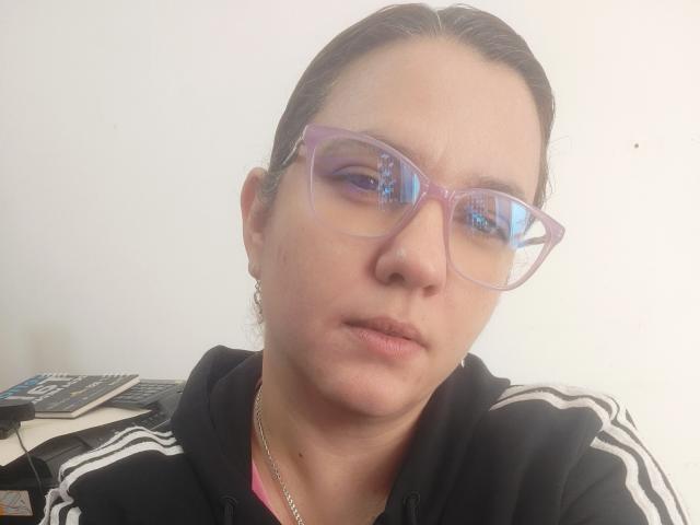 Close-up of AmeliaWilson, wearing clear glasses and a black Adidas hoodie, with a serious expression. She has fair skin, dark hair pulled back, and a silver necklace. Overexposed lighting.