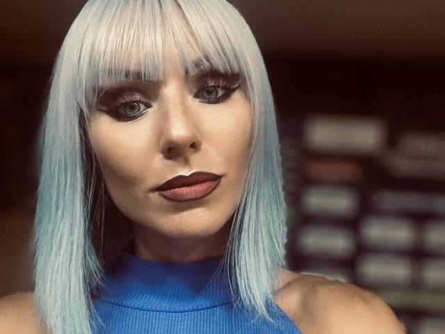 Close-up photo of FortyLeride with straight, silver-blue hair and blunt bangs. She has dark, smoky eye makeup, red lipstick, and wears a sleeveless blue top. Fucking sexy, intense gaze.