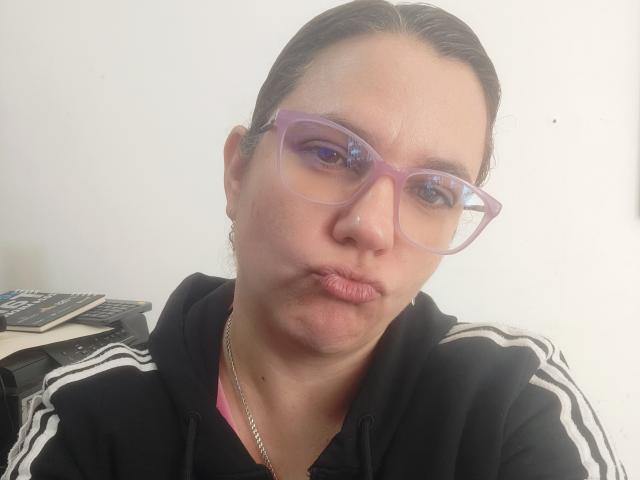 Gros plan d'Amelia Wilson, une femme à la peau brun clair et aux cheveux foncés attachés en arrière, portant des lunettes violettes et un pull à capuche Adidas. Elle fronce les lèvres, créant une ambiance de tension sexuelle..