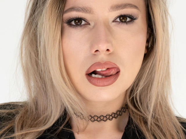 Close-up de flexyHeidi-hot, uma mulher loira com pele clara, lábios cheios e um colar preto. Ela tem olhos castanhos, a boca ligeiramente aberta e uma expressão sedutora. A língua dela está visível..