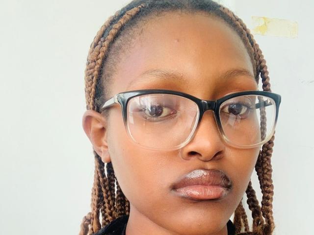 Close-up van YumekoHinata, een Afrikaanse vrouw met donkerbruine huid en gevlochten haar, die een grote bril met zwarte montuur draagt. Kijkend recht in de camera, lippen lichtjes open, intense uitdrukking. Helder licht accentueert haar gezicht..