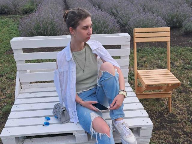 Sanella sits on a white wooden bench with a wooden chair to the right. She's wearing a ripped white shirt, gray top, and blue jeans, with white sneakers. Lavender bushes in the background. Fucking relaxed vibe.