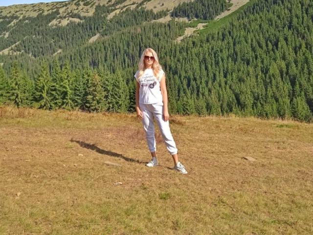 GladisBelle stands in a grassy clearing, wearing a white tank top and pants, sunglasses, and white sneakers. She's facing slightly to the left, with a forested mountain backdrop. Fucking hot.