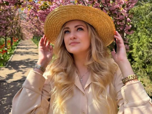 GladisBelle, con lunghi capelli biondi ondulati, indossa un cappello di paglia e una camicetta beige, ed è in piedi su un sentiero soleggiato fiancheggiato da fiori rosa. fiori.
alza lo sguardo con un sorriso appena accennato, con le mani sul cappello..