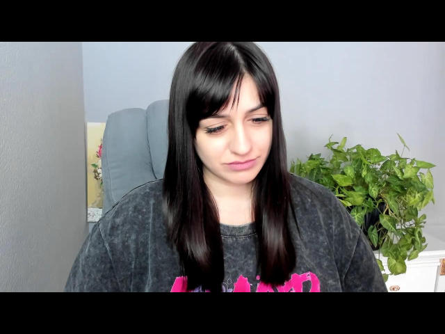 Photograph of a woman with straight black hair, pale skin, and a slight smile. She's wearing a black sweatshirt with pink accents. Green potted plant in the background. Sheidi has a subtle, sexy look.