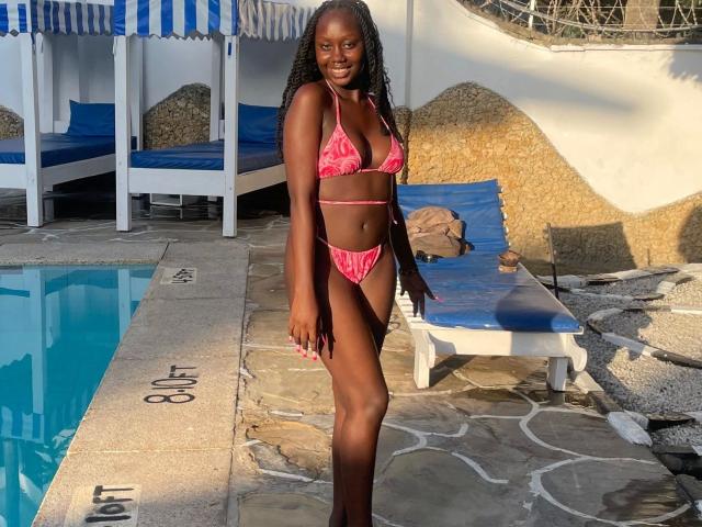SexyDivanah posiert in einem roten Bikini am Pool, ihre dunkle Haut glänzt im Sonnenlicht. Sie hat langes, geflochtenes Haar, kleine Brüste und einen schlanken, attraktiven Körper. Blaue Lounge-Stühle im Hintergrund.