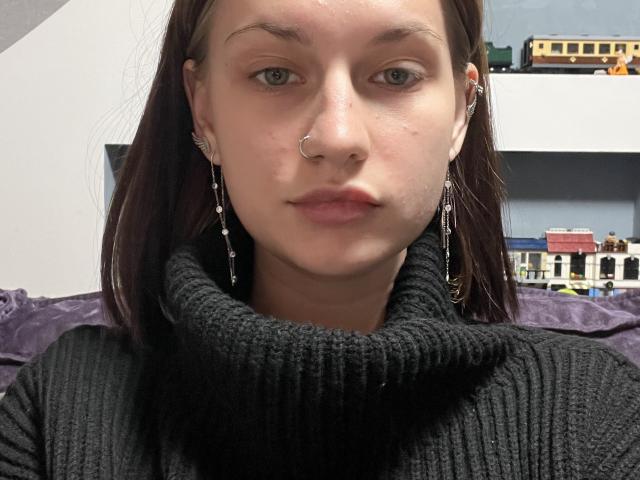 Close-up photo of CleliaMarchak, fair-skinned with dark brown hair, wearing a black turtleneck and silver earrings, nose ring, and hoop earrings, looking directly at the camera, sexual tension present.