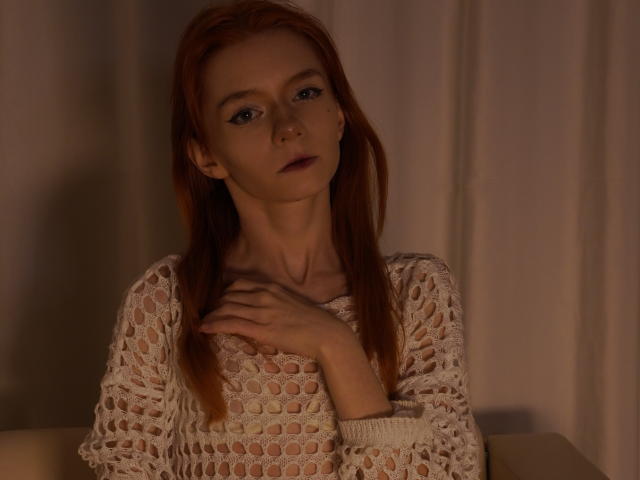 Fotografie von Jamie Lathrum, einer schlanken Frau mit langen roten Haaren, die ein durchsichtiges, weißes Strickoberteil trägt und eine Hand vor die Brust legt. intensiver Blick, gedämpftes Licht, Schatten, die ihre Gesichtszüge betonen, sexuelle Spannung.