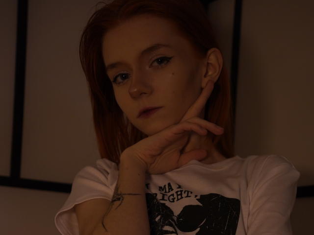 Photographie sombre de JamieLathrum, une jeune femme aux cheveux bruns clairs, aux yeux bleus, et portant un t-shirt blanc avec un imprimé noir. Elle pose son menton sur sa main, et les ombres mettent en valeur son visage. Tension palpable, de nature sexuelle..