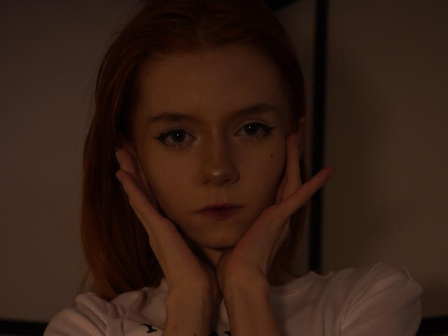Darkly lit photograph of JamieLathrum, hands framing her face, intense blue eyes staring forward, shadows highlighting her features, pale skin, serious expression, slight glint in her eyes, close-up shot.