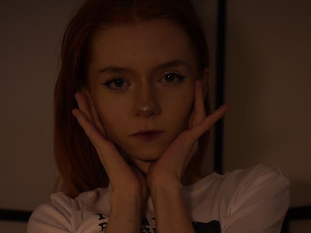 JamieLathrum heeft bleke huid en blauwe ogen, met handen die haar gezicht omlijsten in een foto met weinig licht en donkere tinten. Ze kijkt recht in de camera met een serieuze uitdrukking. Schaduwen accentueren haar gezichtsvorm..